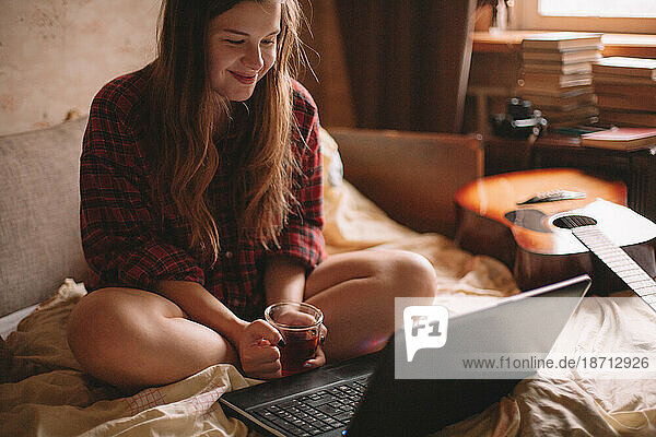 Young woman looking at laptop while having tea sitting on bed