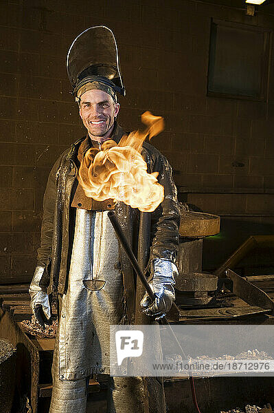 A man in a foudry jacket  coveralls  and a safety helmet with the face guard lifted up holding a flaming tube in one hand and smiling at the camera.