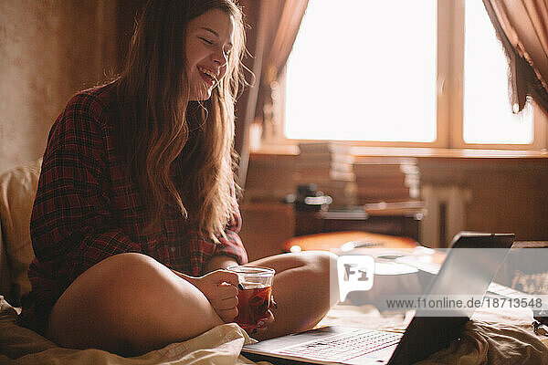 Happy teenage girl with cup of tea sitting in front of laptop