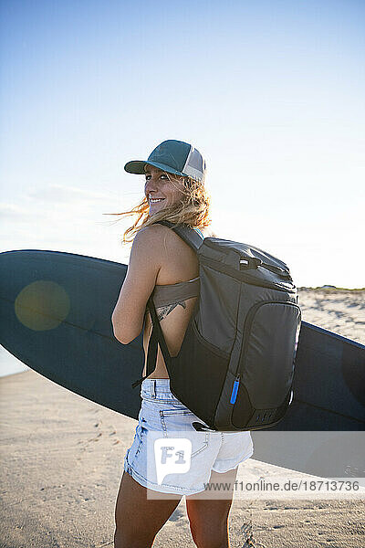 Woman walking the beach at sunset with surfboard and backpack