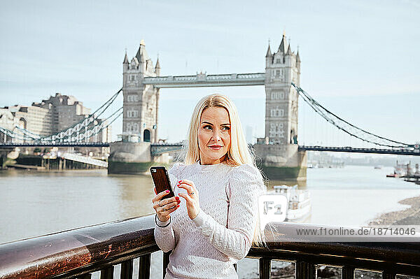 Attractive woman holding mobile phone and looking along on background of Tower Bridge
