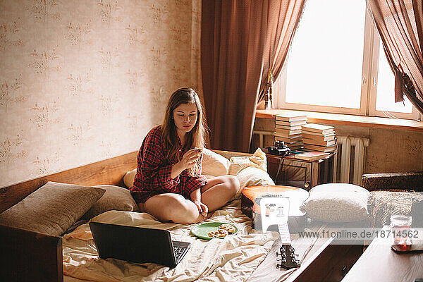 Teenage girl eating in bed looking at laptop