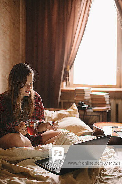 Young woman drinking tea in bed sitting in front of laptop