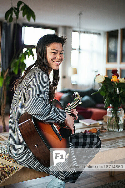 handsome asian woman smiles as she plays guitar in beautiful home