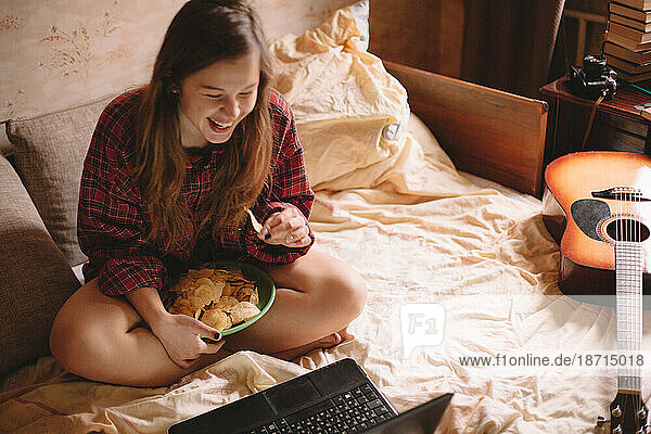 Happy teenage girl eating chips while sitting on bed with laptop