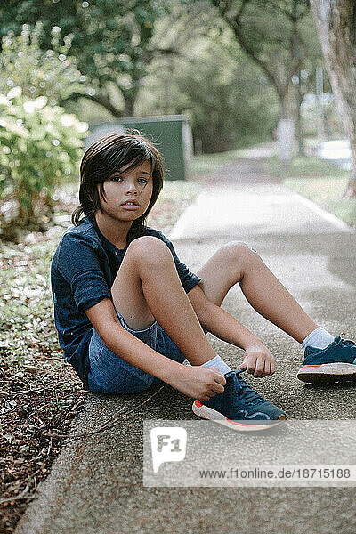 Young multiracial Asian boy sitting on the sidewalk tying his shoes.