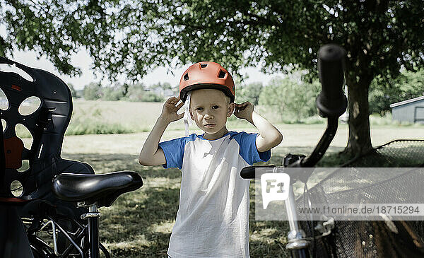 young boy stood next to a bike putting on his helmet