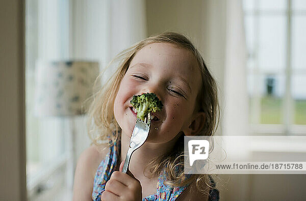 young girl laughing with a messy face whilst eating broccoli
