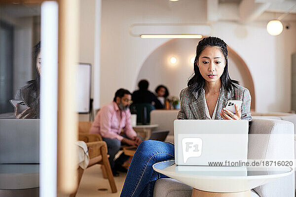Businesswoman holding smart phone using laptop on table