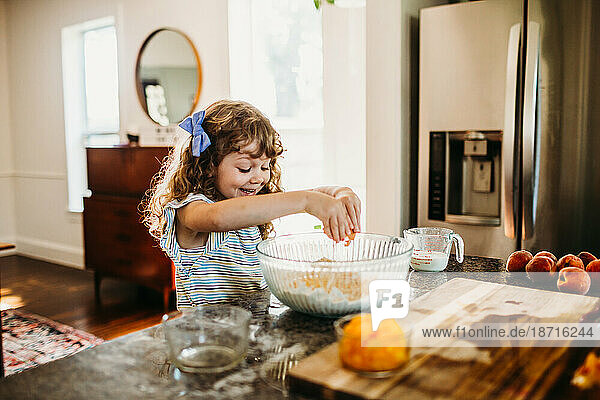 Young girl smiling and cracking raw egg into fresh peach muffin batter