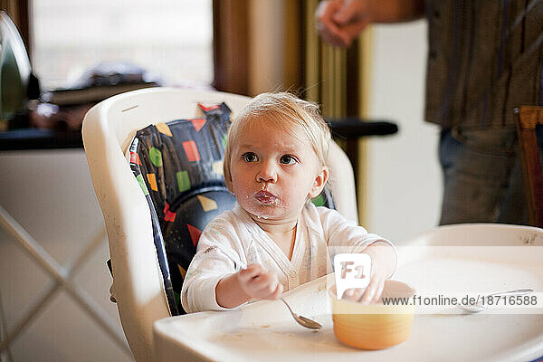 A light haired baby looks up from eating in a high chair in Portland  Oregon.