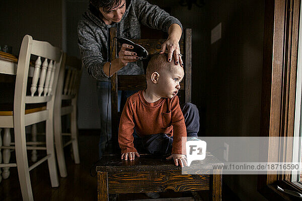 Mom cutting young boys hair at home sitting on a chair looking away