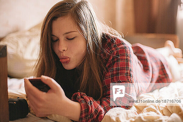 Teenage girl sticking tongue while using smart phone lying on bed