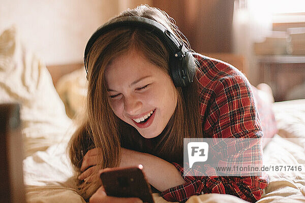 Happy young woman with headphones using smart phone lying on bed