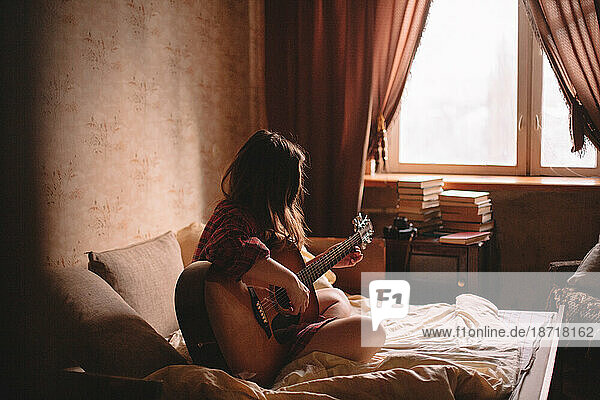 Teenage girl playing guitar while sitting on bed at home