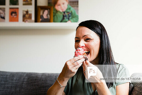 A female laughing while eating a pink cupcake at home