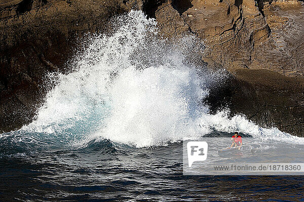 One man paddling an outrigger canoe next to big cliffs and crashing waves.