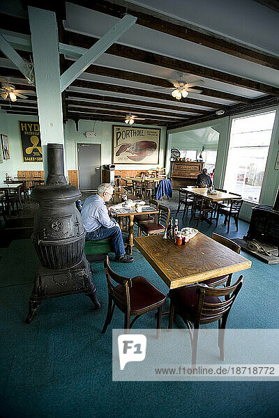 An older gentleman reads the paper by a wood stove in a nearly empty local seafood restaurant in Portland  ME.