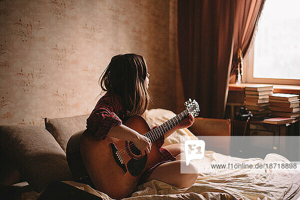 Young woman playing guitar while sitting on bed at home