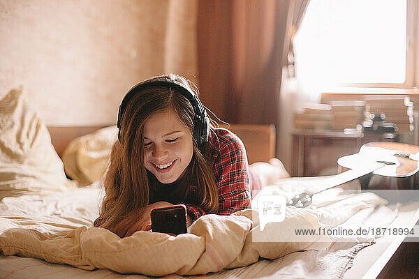 Happy teenage girl with headphones using smartphone lying on bed