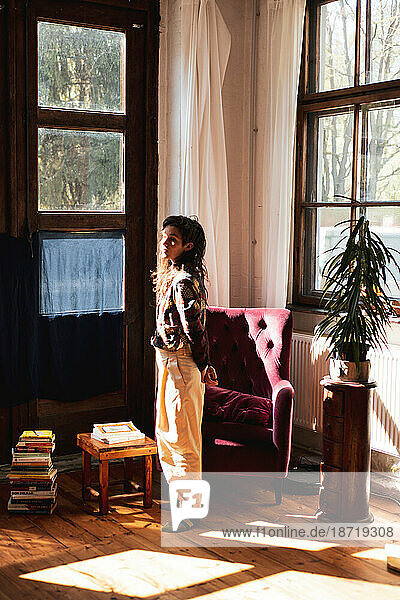 woman stands in large window light at home with plants and books