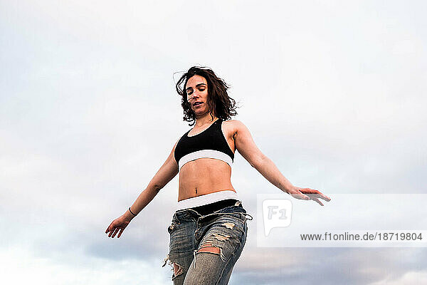 Young woman balances with sky background wearing sports bra and jeans