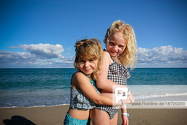 two girls hugging standing on the beach in bathing suits on sunny day