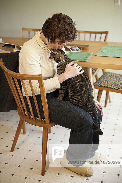 A woman kisses a tabby cat she is holding on her lap in a kitchen in Seattle  Washington.