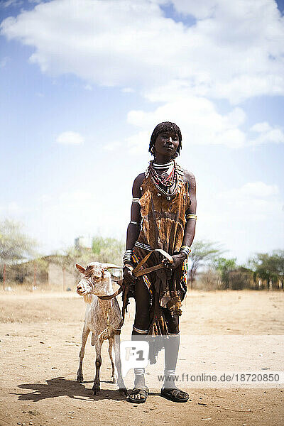 A portrait of a woman standing next to her goat. She is dressed in traditional clothes of the Hamer people.