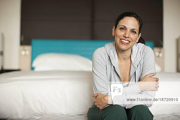 A hispanic woman wearing a sweatshirt sits in her bedroom at the foot of the bed and looks on camera.