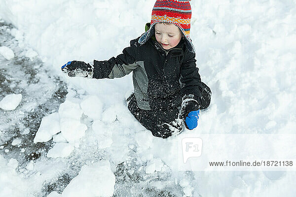Young boy playfully kneels in snow making snowballs after snowstorm