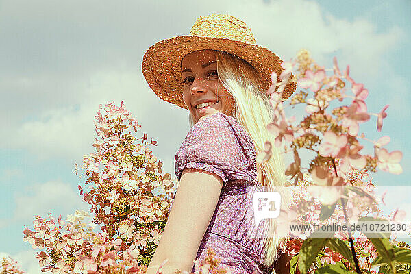 Smiling young blonde woman in a straw hat near hydrangea flowers