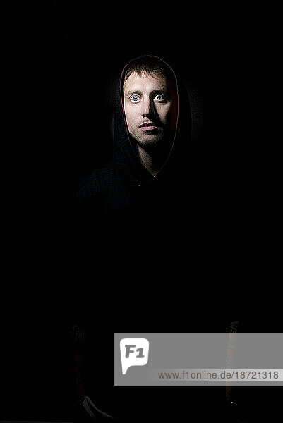 Portrait of young man with blue eyes and a hood on with a scared facial expression on black backdrop