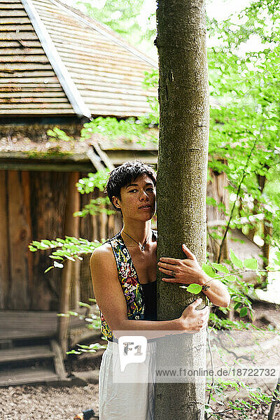 strong androgynous asian woman embraces tree by wooden house