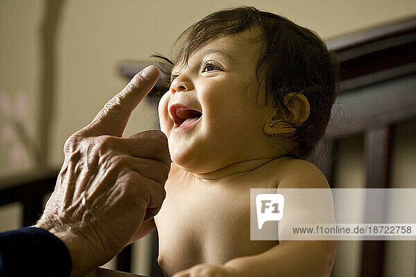 An adorable baby girl giggles as she is patted on the nose while holding herself up in her crib.