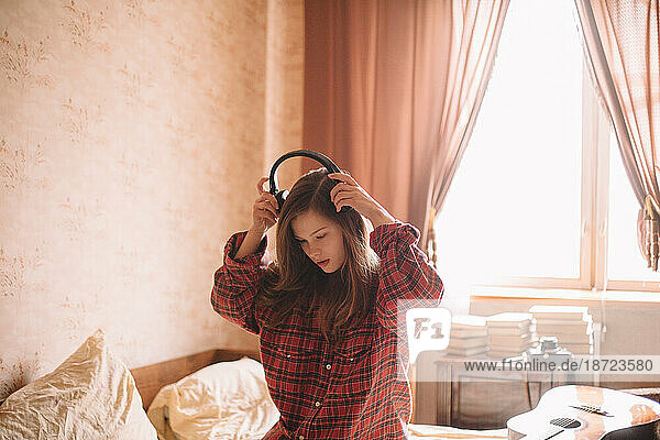Young woman putting on headphones while sitting on bed at home