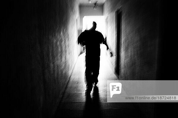 A gypsy man walks down a long hallway  silhouetted by the light at the end  in the converted cow-house where has lived with his family in exile for two years
