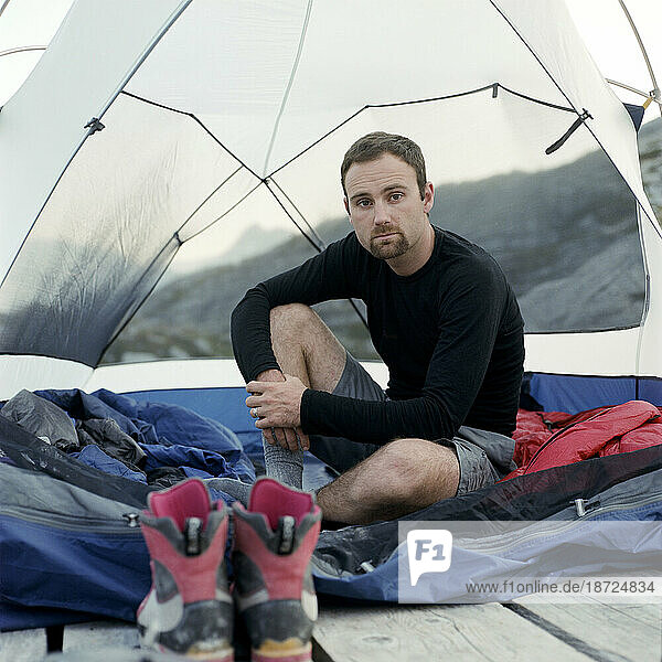 A man poses for a portrait inside of a tent pitched in the mountains.