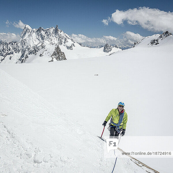 A smiling mountaineer approaches a steep snow slope on a bluebird day