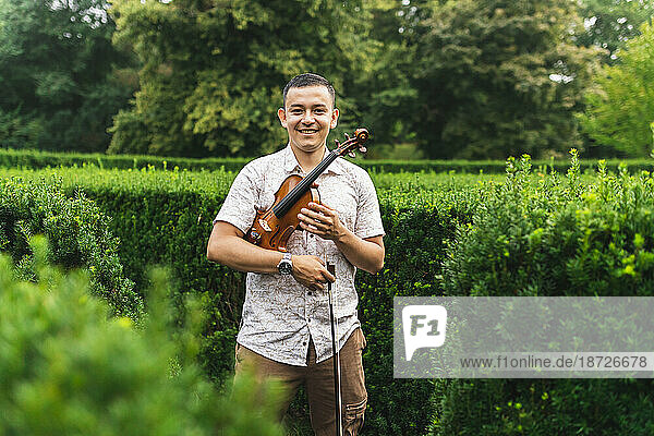 Smiling man holding violin and bow standing in front of plants at park