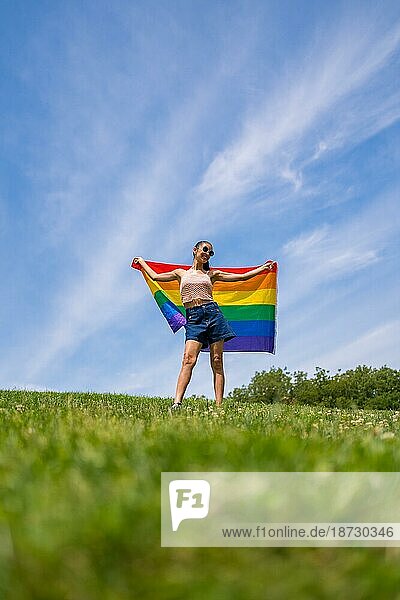 Caucasian brunette Frau mit einem Regenbogen lgbt Flagge auf dem Gras und blauen Himmel Hintergrund