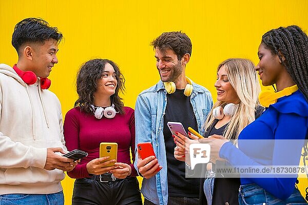 Niedriger Blickwinkel auf eine Gruppe lächelnder junger multiethnischer Teenager-Freunde  die Mobiltelefone auf einem gelben Hintergrund benutzen. Technologie süchtig Millennial Gemeinschaft Konzept. Social Media Kommunikation Generation Z