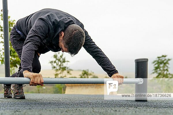 Junger  bärtiger  schwarz gekleideter Mann  der an einem regnerischen Tag in einem Fitnessstudio im Freien mit einer Hantel auf dem Boden trainiert und Dehnübungen macht