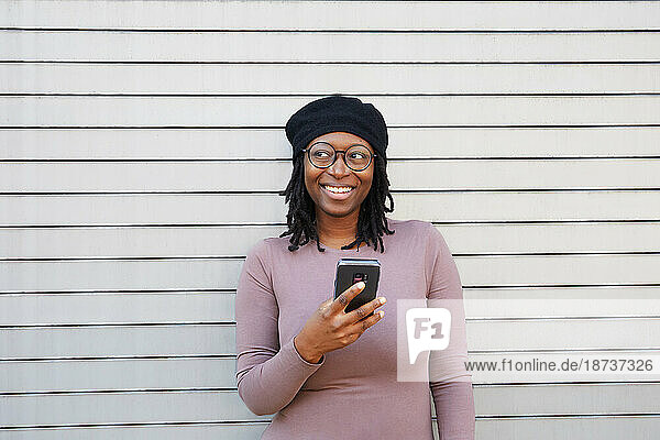 Smiling woman wearing eyeglasses and beret holding smart phone in city