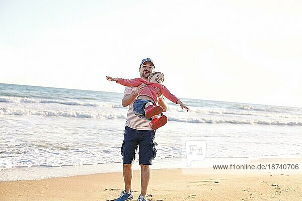 Liebevoller Vater spielt mit seiner entzückenden kleinen Tochter am Strand  schaukelt sie herum  Vater und Kind verbringen Zeit im Freien zusammen  sonniges Meer im Hintergrund. Parenting Konzept
