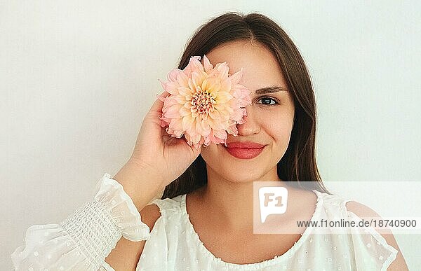 Fröhliche junge Frau schaut lächelnd in die Kamera und bedeckt ihr Auge mit einer natürlichen Blume vor weißem Hintergrund