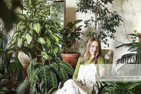 Ruhige Frau  die auf einem Stuhl in einem Raum voller Blumentöpfe mit üppigen Pflanzen faulenzt