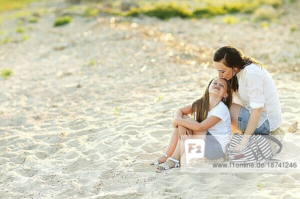 Liebevolle Mutter küsst bezauberndes Mädchen auf die Stirn  während sie im Sommer bei Sonnenuntergang zusammen am Ufer sitzen