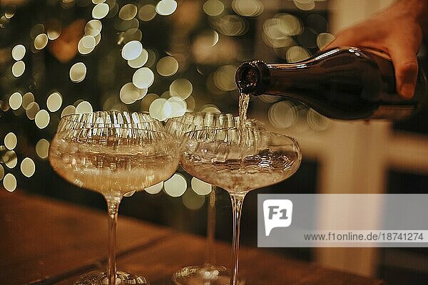 Silvesterfeier. Mann gießt Champagner in Gläser stehen auf dem Tisch mit festlichen Weihnachtsessen  Kerzen und eingepackt Geschenke gegen unscharfen Hintergrund und firtree  beschnitten