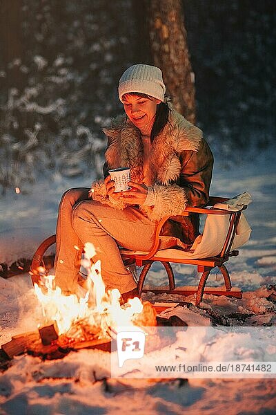 Glückliche junge Frau voller Ruhe und Gelassenheit in warmer Strickmütze und Jacke sitzt auf einem Holzschlitten am Lagerfeuer mit einer Tasse heißem Tee in der Hand  umgeben von einem schneebedeckten Winterwald